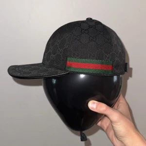 Svart keps från Gucci med monogram - Svart keps från Gucci med diskret GG-monogram över hela kepsen. Klassisk böjd skärm och grönt/rött band på sidan. Kepsen har en stilren och lyxig känsla, perfekt för dig som gillar designeraccessoarer. Har också S och L om M inte passar just dig. 
