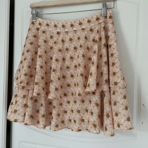 Blommig volangkjol från NA-KD - Supersöt kort kjol med volanger från The Fashion Fraction x NA-KD. Kjolen är beige med blommigt mönster i vitt och brunt, har en flowy passform och är perfekt för varma dagar. Använd en gång.