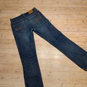 Blå jeans från Nudie Jeans - Säljer ett par klassiska blå jeans från Nudie Jeans med snygga orange sömmar och broderad bakficka. Modellen har normal passform och raka ben. Perfekta för dig som gillar stilrena och tidlösa jeans. Storlek W32L34