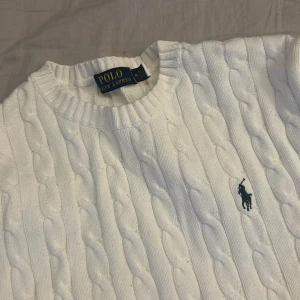 Klassisk Vit kabelstickad tröja från Polo Ralph Lauren - Vit kabelstickad tröja från Polo Ralph Lauren med rund halsringning och blå broderad logga på bröstet. Inga defekter och är i nyskick!!!Klassisk och stilren passar mycket bra nu inför sommaren inte äkta!!!.