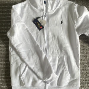 Vit zip hoodie från Polo Ralph Lauren - Säljer en klassisk vit hoodie från Polo Ralph Lauren med dragkedja och fickor framtill. Tröjan har det ikoniska broderade logotypen på bröstet och är tillverkad i mjukt material. Digitalt kvitto finns även.