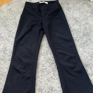 Svarta kostymbyxor från Bershka - Snygga svarta kostymbyxor från Bershka med klassisk passform och raka ben. Perfekta för en stilren och elegant look. Byxorna har hällor för bälte och stängs med knapp och dragkedja framtill.