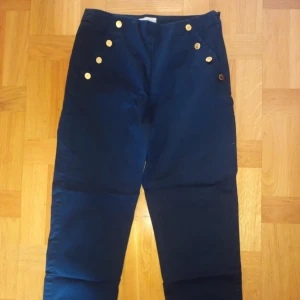 Marinblå byxor med guldknappar från Holly & Whyte - Snygga marinblå byxor från Holly & Whyte med dekorativa guldknappar framtill. Byxorna har en klassisk rak passform och är perfekta för en stilren look. Tillverkade i ett mjukt bomullsmaterial.