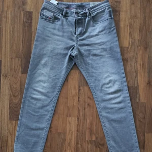 Grå jeansbyxor från Diesel Buster W34 L30 - Säljer ett par grå jeansbyxor från Diesel, modell Buster. Byxorna har klassisk femficksdesign, normal passform och raka ben. Snygga röda sömmar på insidan ger en extra detalj. Perfekta för dig som gillar stilrena och bekväma jeans.