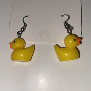 Gula örhängen med bad anka-motiv INGA PLUPPAR - Supergulliga örhängen med små gula ankor i plast. Perfekta för dig som vill sticka ut och addera lite lekfullhet till din stil. Hängande modell med krok i metall och detaljer i orange och svart.