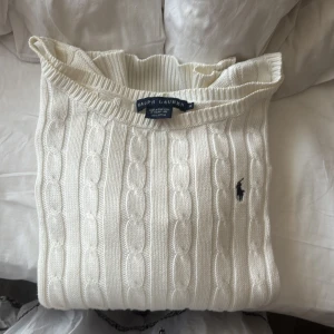 Vit kabelstickad tröja från Ralph Lauren - Säljer en klassisk vit kabelstickad tröja från Ralph Lauren med rund halsringning och diskret broderad logga på bröstet. Tröjan har långa ärmar och ribbade muddar vid ärmslut och nederkant. Perfekt för en stilren look.