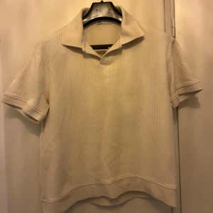 Beige ribbad pikétröja - Säljer en stilren beige pikétröja med korta ärmar och klassisk krage. Tröjan är ribbad och har en avslappnad passform, perfekt för en enkel och snygg look. Strl M, men sitter som en S