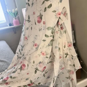 Vit blommig långklänning med rosett - Romantisk långklänning i vitt med rosa blommor och gröna blad. Klänningen har axelbandslös design med en söt rosett på ena axeln och volangdetaljer längs kjolen. Perfekt för sommarens speciella tillfällen.