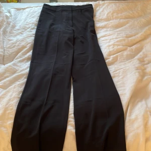 Svarta kostymbyxor från H&M - Säljer ett par svarta kostymbyxor från H&M med klassisk rak passform och pressveck. Byxorna har hög midja och vida ben, perfekta för en stilren look.