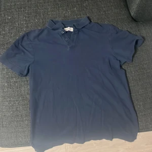 Mörkblå pikétröja från These Glory Days - Säljer en stilren mörkblå pikétröja från These Glory Days. Tröjan har klassisk krage och knappar framtill, perfekt för en avslappnad men snygg look. Passar dig som gillar enkel och tidlös stil.