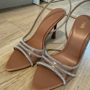 Beige klackskor med transparenta band och strass från Zara - Säljer ett par snygga beige klackskor från Zara med transparenta band dekorerade med strass. Skorna har öppen tå och smal klack, perfekta för att lyfta din outfit. Använda en gång.