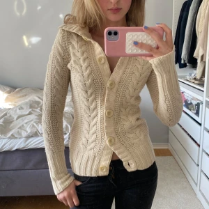 Beige stickad kofta från Madison Lilly - Säljer en beige stickad kofta från Madison Lilly med kabelstickat mönster och stora knappar framtill. Koftan har lång ärm, bred krage och ribbade muddar. Perfekt för lager-på-lager och en mysig stil.