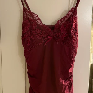 Vinrött spetslinne - Säljer ett linne med vacker spetsdetalj upptill och en liten rosett framtill. Linnet har smala axelband och är perfekt för en festlig kväll. Materialet är mjukt och bekvämt, vilket gör det både stiligt och skönt att bära.