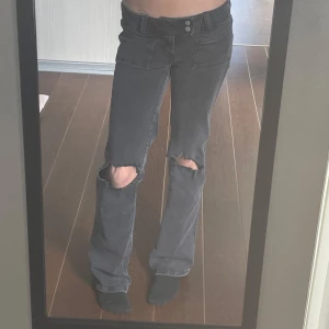 Grå bootcut jeans med hål på knäna - Säljer ett par gråa bootcut jeans med slitna hål på båda knäna. Jeansen har låg midja, klassiska fickor och stängs med dragkedja och dubbla knappar. 