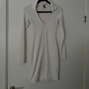 Vit ribbad klänning med dragkedja - Säljer en vit ribbad klänning med lång ärm och djup v-ringning. Klänningen har en dragkedja framtill och en bred krage som ger en cool look. Perfekt för dig som gillar stilrena plagg med lite edge.