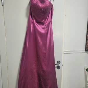 Mörkrosa långklänning från Juju & Christine - Säljer en elegant mörkrosa långklänning från Juju & Christine. Klänningen är axelbandslös med glittriga detaljer framtill och har snörning i ryggen för en justerbar passform. Perfekt för bal eller festliga tillfällen.