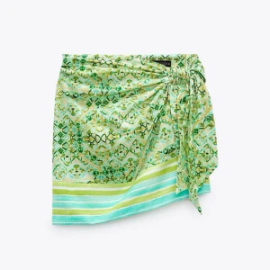 Grön omlottkjol med mönster från Zara - Säljer en kort grön omlottkjol från Zara med knyt i sidan. Kjolen har ett färgglatt mönster i grönt, gult och turkost samt ränder längst ner. Toppen skick även lite liten i storleken så skulle klassa den som en XS🩵