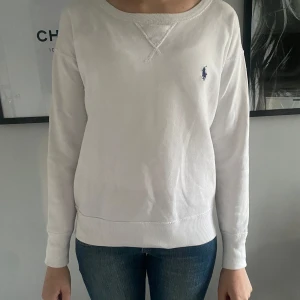 Vit sweatshirt från Ralph Lauren - Säljer en klassisk vit sweatshirt från Ralph Lauren med rund halsringning och broderad blå logga på bröstet. Tröjan har långa ärmar och ribbade muddar vid ärmslut och nederkant. Perfekt för en stilren och avslappnad look.