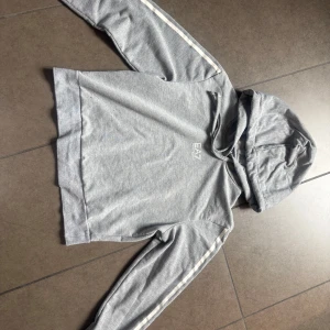 Grå hoodie från Emporio Armani EA7 - Säljer en stilren grå hoodie från Emporio Armani EA7. Tröjan har huva och långa ärmar med vita ränder och logga längs ärmarna. Perfekt för en avslappnad look och enkel att matcha med det mesta. Enbart använd 2 gånger. Nypris: 1 300kr