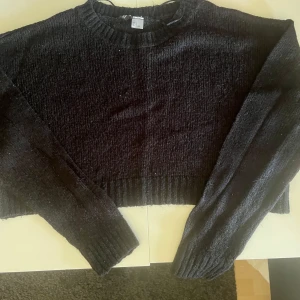 Svart croppad stickad tröja - Säljer en svart croppad stickad tröja med rund halsringning och långa ärmar. Perfekt att matcha med höga jeans eller kjol. Tröjan har ribbade muddar vid ärmslut och nederkant för en snygg passform.