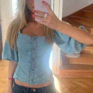 Ljusblå jeanstopp med puffärmar - Säljer en ljusblå korsettopp i jeansmaterial med puffiga korta ärmar och knappar framtill. Toppen har en fyrkantig urringning och figurnära passform. Perfekt för dig som vill ha en trendig och snygg topp till sommaren!