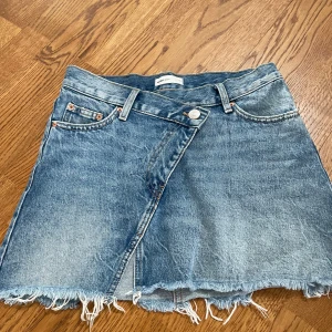 Jeanskjol från Perfect Jeans - Säljer en snygg blå jeanskjol från Perfect Jeans med omlott-look och råa kanter nertill. Kjolen har klassiska fickor fram och bak samt bälteshällor. Perfekt för dig som gillar en avslappnad och trendig stil.