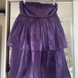 Axelbandslös volangklänning från NA-KD - Llila axelbandslös klänning från NA-KD med volanger och glansigt material. Helt ny med lappen kvar passar perfekt nu till sommaren och festliga tillfällen. 