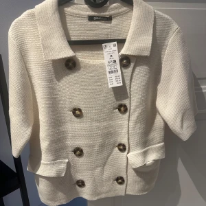 Vit cardigan med knappar från Gina Tricot - Vit cardigan från Gina Tricot med knappar framtill och två fickor. Jackan har trekvartsärm och en krage. Har aldrig kommit till användning och har prislappen kvar. 