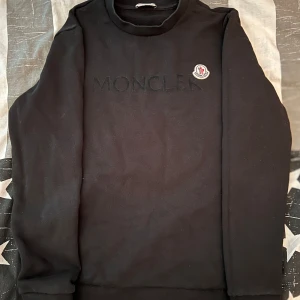Moncler Tröja - Säljer en svart sweatshirt från Moncler med broderad logotyp på bröstet och Moncler-tryck framtill. Tröjan har rund halsringning och långa ärmar. Perfekt för en stilren och avslappnad look.