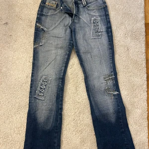 Blå jeansbyxor  från Jeans yyp - Säljer ett par blå jeansbyxor från Jeans yyp med slitningar och snörningsdetaljer på både fram- och bakfickor. Byxorna har raka ben och en cool, tvättad look med ljusare partier. Perfekta för dig som gillar en unik och edgy stil.det finns en fläck som syns på sista bilden 