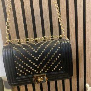 Svart axelväska med nitar och guldkedja - Säljer en svart axelväska med guldfärgade nitar i mönster och en kraftig guldkedja som axelrem. Väskan har ett snyggt lås framtill och är tillverkad i syntetmaterial. Perfekt för dig som vill ha en cool accessoar med attityd.