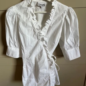Vit omlottblus med volang från Ganni - Säljer en vit omlottblus från Ganni med volangdetaljer längs v-ringningen och knyt i sidan. Blusen har korta puffärmar och är tillverkad i ett lätt bomullsmaterial. Perfekt för vår och sommar!