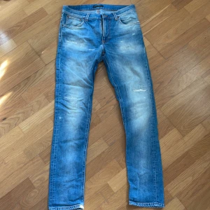 Blå jeans från Nudie Jeans - Säljer ett par klassiska blå jeans från Nudie Jeans med slitningar och ljusa partier på benen. Jeansen har fem fickor, orangea sömmar och rak passform. Perfekta för en avslappnad stil.