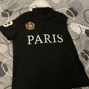 Svart Paris t-shirt med emblem - Svart t-shirt med trycket 'PARIS' på bröstet, stort emblem med krona och amerikansk sköld, samt siffran 3 i vitt och gult på ena ärmen. Klassisk krage med knappar framtill. Perfekt för dig som gillar sportig och urban stil.