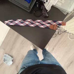 Onewear bälte  - Säljer ett snyggt flätat bälte med randigt mönster i blått, vitt och rött. Bältet har bruna läderdetaljer vid spännet och ändarna, vilket ger en schysst kontrast. Perfekt accessoar för att lyfta din outfit. 