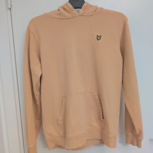 Beige hoodie från Lyle & Scott 170/ S - Snygg diskret korall/orange hoodie från Lyle & Scott med klassisk gul logga på bröstet. Tröjan har huva, känguruficka och ribbade muddar vid ärmslut och nederkant. Strl motsvarar 170 i barn. S i herr