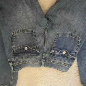 Blå jeans från Gina Tricot med fickdetaljer - Säljer ett par klassiska blå jeans från Gina Tricot i storlek 158. Jeansen har snygga fickor med lock och knapp bak, samt bälteshällor och knappgylf. Perfekta för dig som gillar en tidlös denimlook.