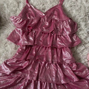 Glittrig rosa volangklänning  - Supersöt rosa klänning med glittrigt tyg och flera lager volanger. Klänningen har smala axelband och en v-ringad front. Perfekt för dig som vill sticka ut med en färgstark och glammig look. Materialet är polyester och klänningen är kort.