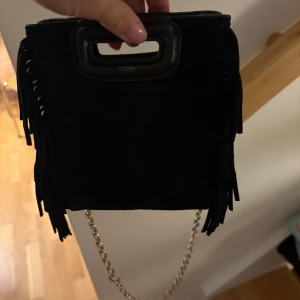 Svart mocka axelväska med fransar från Maje - Snygg svart axelväska i mocka från Maje med coola fransdetaljer på sidorna och guldfärgad kedja. Tillkommer ett svart band som är lite längre. Äkthetsbevis medföljer och dustbag