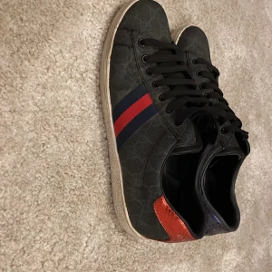 Mörkblå Gucci ace skor - Snygga svarta Gucci sneakers med klassiskt GG-mönster, röda och marinblå ränder på sidorna och detaljer i rött och blåt på hälen. Skorna har svarta skosnören och vit sula. Kommer med originalkartong och dustbag. Skorna är mycket fint skick och har endast två små slitningar på under sulan. 