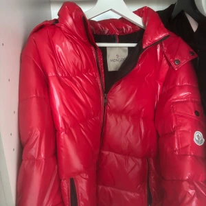 Röd pufferjacka från Moncler - Säljer en riktigt snygg röd pufferjacka från Moncler. Jackan har glansig yta, huva, dragkedja och fickor med dragkedja samt en ärmficka med Moncler-logga. Perfekt för kalla dagar och ger en cool streetstil. size S-M