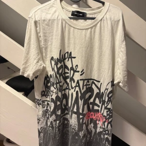 Vit t-shirt med graffiti från Dsquared2 - Dsquared 2 T-shirt vit i storlek XXL med coolt svart och rosa graffitimönster över hela framsidan och baksidan. T-shirten har rund halsringning och är kortärmad. Materialet är mjuk bomull som känns skön mot huden.