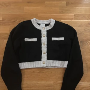 Svart och vit croppad kofta med pärlknappar - Snygg croppad kofta från H&M i svart med vita detaljer och pärlknappar framtill. Koftan har långa ärmar, ribbade muddar och två dekorativa fickor på bröstet. Perfekt för dig som gillar en stilren och trendig look.