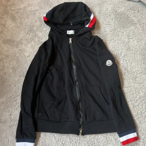 Svart hoodie från Moncler med dragkedja - Svart hoodie från Moncler med hel dragkedja, huva och Moncler-logga på ärmen. Röd, vit och blå rand på huva och ärmslut ger en sportig vibe. Perfekt för dig som gillar stilrena och bekväma plagg. Använd 1-3 gånger 