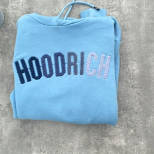 Blå HOODRICH hoodie med brodyr - Snygg ljusblå hoodie från HOODRICH med broderad logga i olika blå nyanser över bröstet. Klassisk känguruficka, huva med dragsko och mjukt material som känns skönt mot huden. Perfekt för chill dagar eller streetwear-stil.