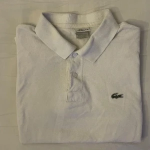 Vit pikétröja från Lacoste - Snygg vit pikétröja från Lacoste med klassisk krage och två knappar framtill. Tröjan har det ikoniska gröna krokodilmärket broderat på bröstet. Tillverkad i mjuk bomull som andas och passar perfekt till en chill stil.