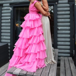 Rosa långklänning med volanger - Säljer en maxad rosa långklänning med flera lager volanger och djup v-ringning. Klänningen har breda axelband och markerad midja, vilket ger en riktigt snygg siluett. Perfekt för bal eller bröllop! Materialet är lätt och ger ett härligt flow när du rör dig. Det finns bara en sådan klänning då den är omsydd för hur jag ville att den skulle se ut! Den är köpt för 7000kr