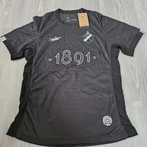 Svart AIK Smokinglirare fotbollströja Nike - AIK Smokinglirare Edition 2025 fotbollströja från Nike i svart med subtila vertikala ränder. Tryck med '1891' på bröstet, AIK-emblem och Smokinglirare-märke på ryggen. Kortärmad, lätt och ventilerande material perfekt för match eller träning.