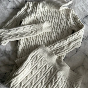 Vit stickad vinter klänning  - Vinter klänning med hög krage och långa ärmar. Klänningen  har klassiskt flätmönster och breda ribbade muddar vid ärmslut och nederkant. 