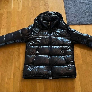 Svart Moncler pufferjacka med huva - Säljer en svart, glansig pufferjacka från Moncler med huva och dragkedja framtill. Jackan har två snedställda fickor med dragkedja och en liten ficka med Moncler-logga på bröstet. Perfekt för kalla dagar när du vill vara både varm och snygg. Storlek L. Använd en fåtal gånger, nästan som ny. 